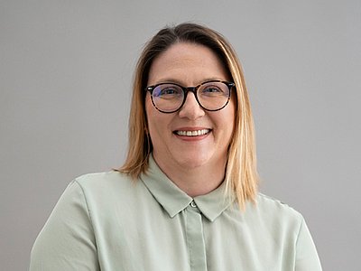 Iris Schweiger trägt blode halblange Haare, eine dunkle Brille, eine hellgrüne Bluse und lächelt in die Kamera.