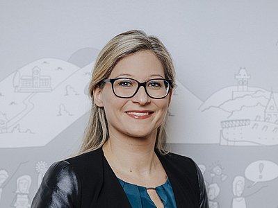 Simone Kosnik trägt einen schwarzen, glänzenden Blazer und eine dunkelblaue Bluse. Ihr langes, blondes Haar ist am Hinterkopf teilweise zusammengebunden. Sie trägt eine Brille mit dunklen Rahmen und lächelt in die Kamera.
