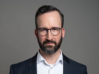 Maximilian Brandl blickt die Betrachtenden an. Er trägt einen Vollbart und eine Brille. Unter seinem dunklen Blazer trägt er ein helles Hemd.