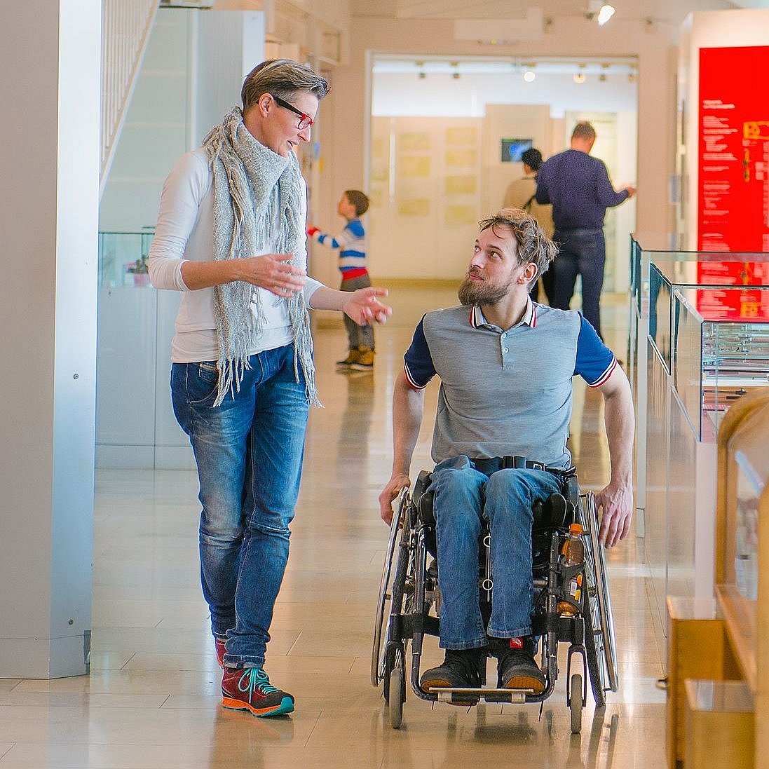 Ein Vorstellungsgespräch oder erster Arbeitstag: Zwei Personen befinden sich im Gespräch, in einem Flur. Beide sind in Bewegung: eine Person geht und eine bewegt sich mittels Rollstuhls fort.