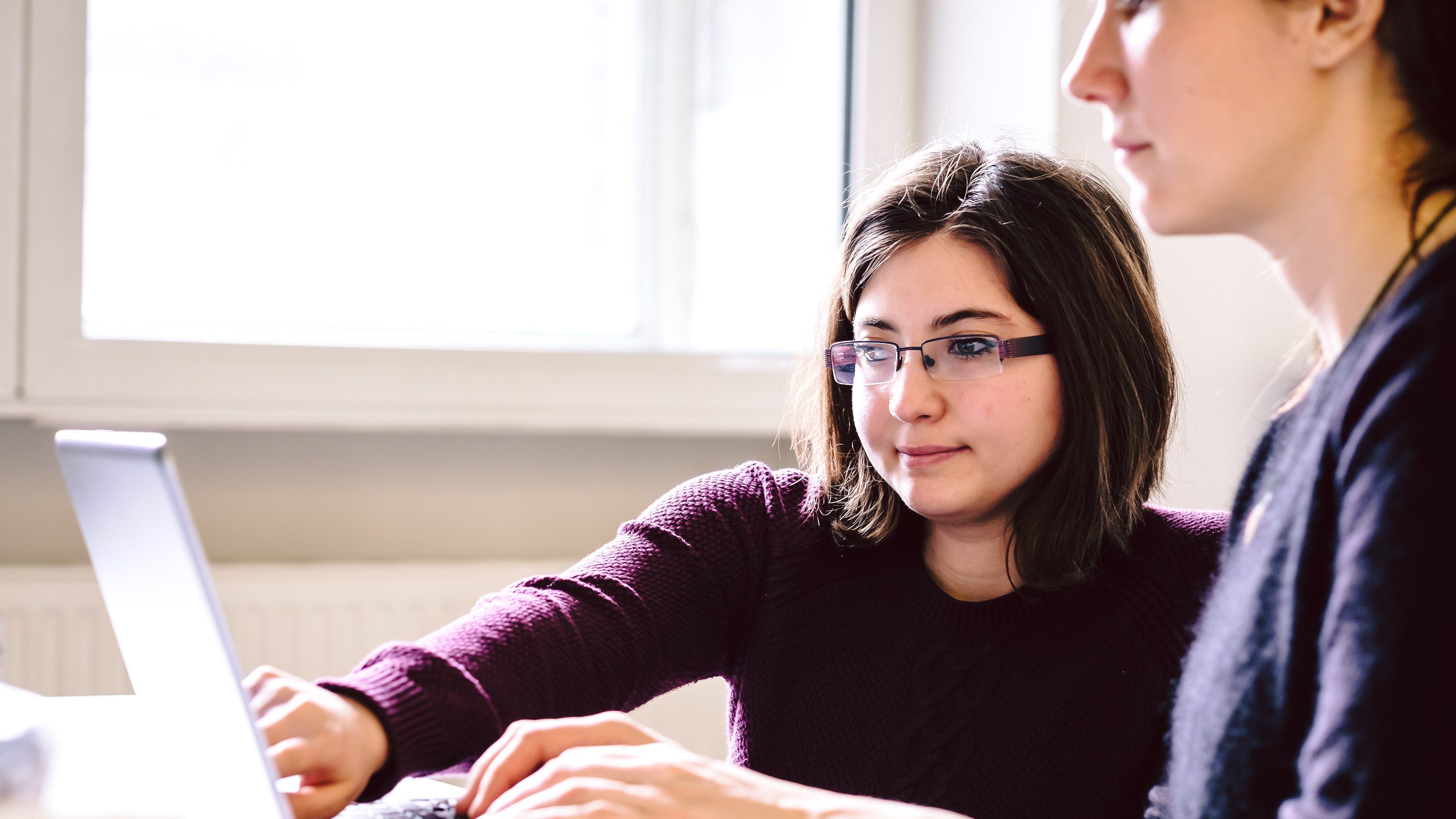Büro-Situation: Zwei Personen, die als Frauen und unterschiedlicher Herkunft gelesen werden können, arbeiten gemeinsam an einem Schreibtisch, nebeneinandersitzend. Während eine von beiden auf den Bildschirm des Laptops zeigt, tippt die andere auf der Tastatur. Die schreibende Frau tippt mit geschlossenen Augen.